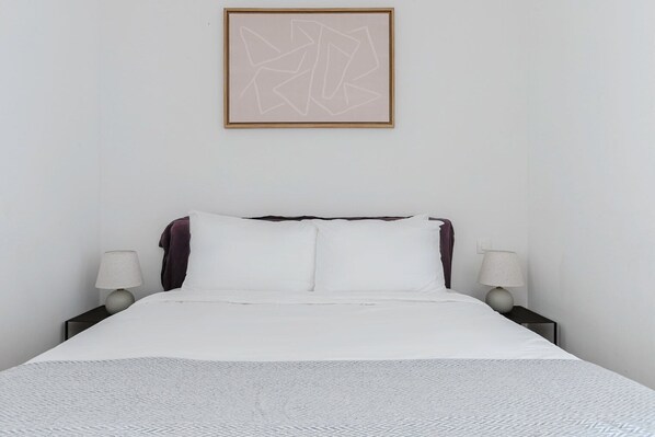 Una cama bien hecha con sábanas blancas, dos lámparas de noche y una obra de arte abstracta enmarcada en la pared.