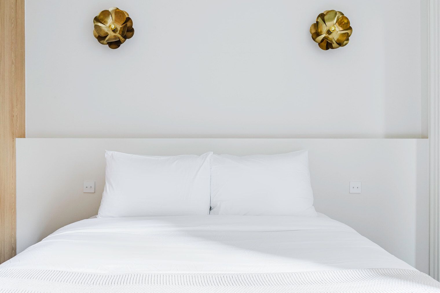 A neatly made bed with white bedding, a wooden headboard, and two wall-mounted decorative gold fixtures.