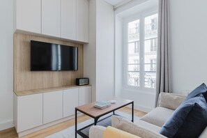 Un salon moderne avec un téléviseur à écran plat fixé sur un mur recouvert de panneaux de bois, une petite table avec des livres et une radio, un canapé avec des coussins et une grande fenêtre donnant sur des immeubles.