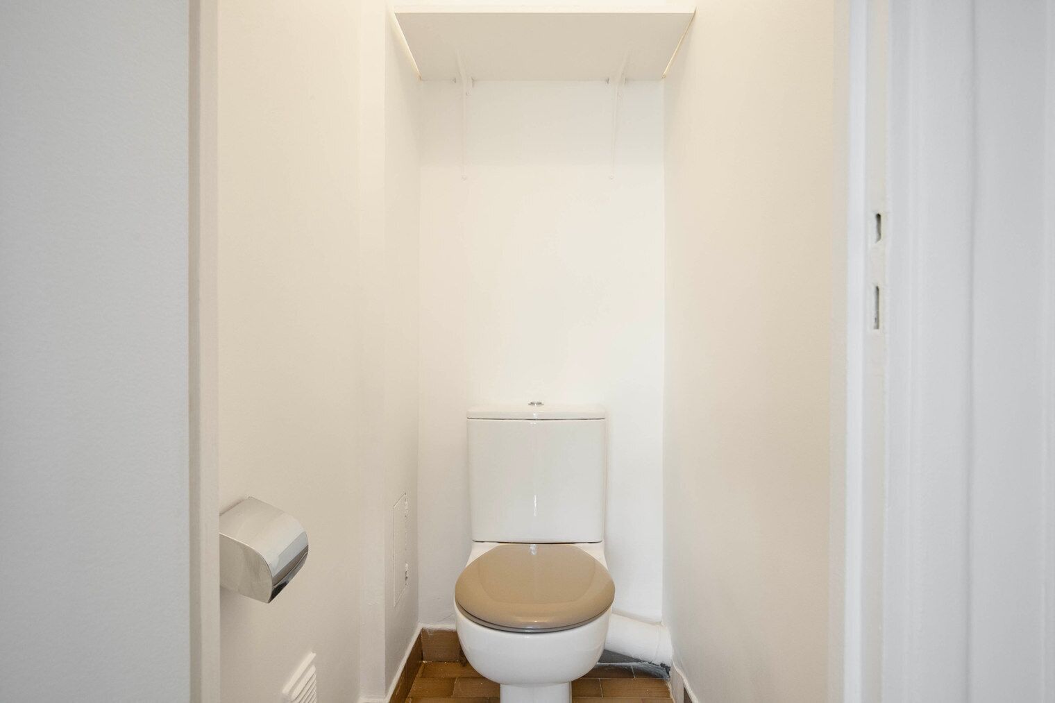 A small, narrow bathroom with a toilet, a toilet paper holder, and a wooden floor.