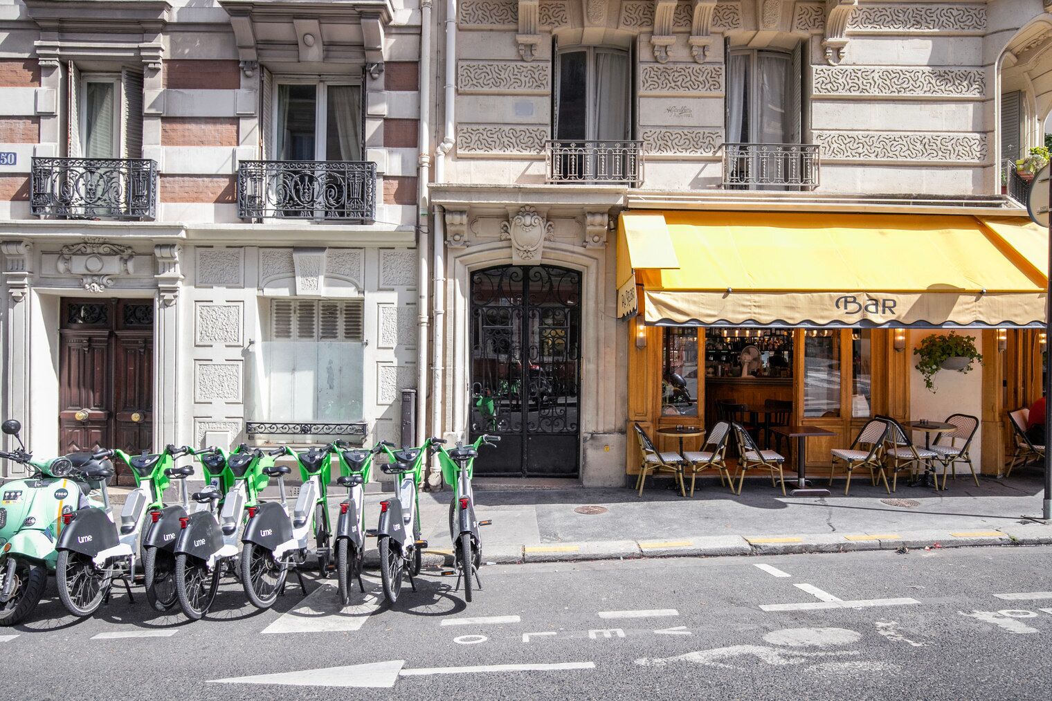 Een rij elektrische Lime-scooters geparkeerd voor een gebouw met een geel luifel waarop 'Bar' staat.