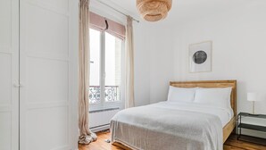 A bedroom with a wooden bed, white bedding, a hanging light fixture, a radiator, and a window with curtains.