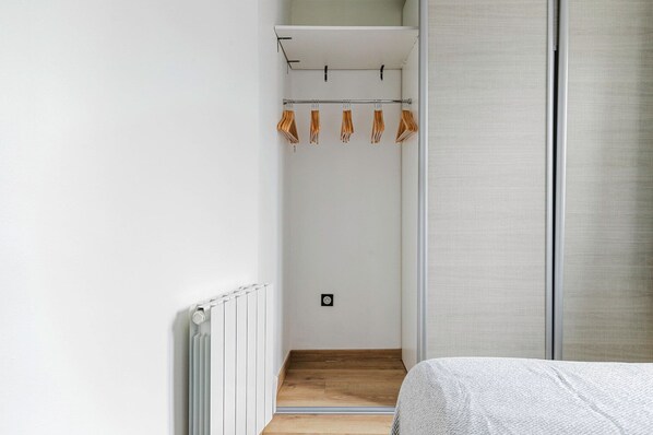 Un pasillo estrecho con un radiador, suelo de madera y una cama con cubierta blanca texturizada.