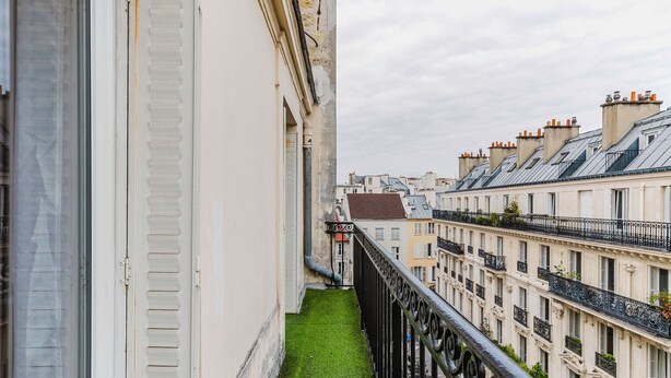 Ein schmaler Balkon mit einem kleinen Rasenstück, mit Blick auf eine Reihung traditioneller europäischer Gebäude mit Balkonen und Schornsteinen.