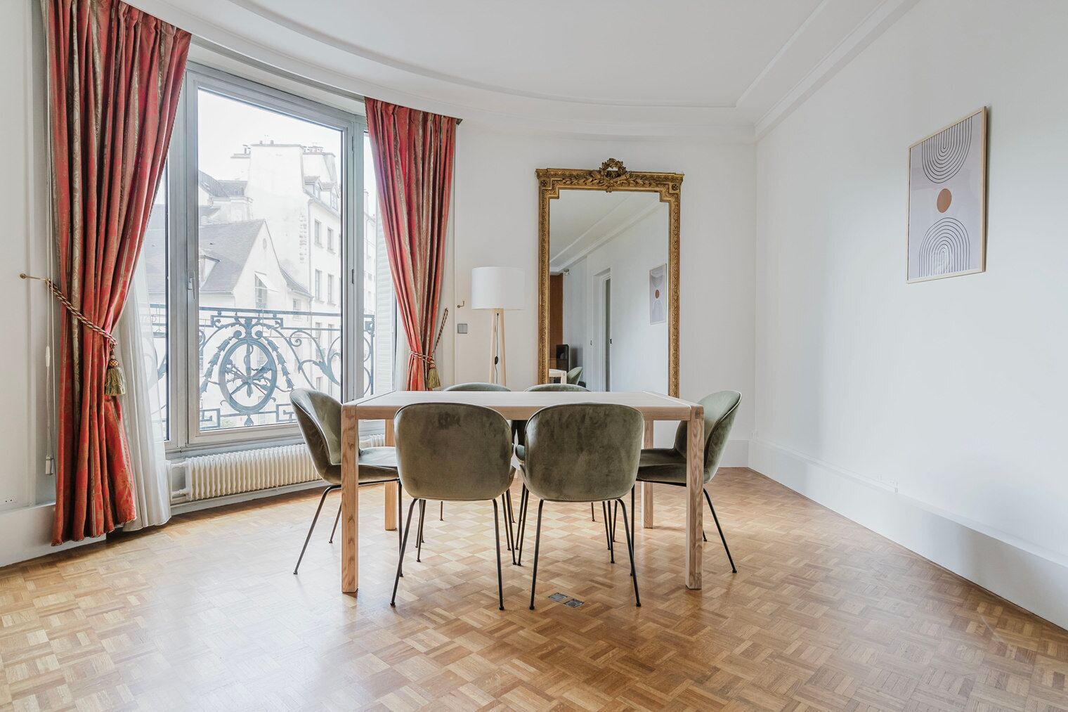 Een eetruimte met een houten tafel en vier stoelen, een grote spiegel en uitzicht op gebouwen door een raam met rode gordijnen.