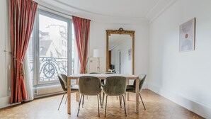A dining area with a wooden table and four chairs, a large mirror, and a view of buildings through a window with red curtains.