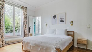 A bedroom with a large bed, a wooden nightstand, a radiator, and a view of greenery through the window.