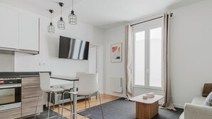 A modern kitchen with a built-in oven, a dining area with a table and chairs, a large window with curtains, and a flat-screen TV mounted on the wall.