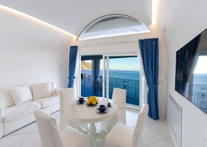 Une pièce moderne avec un canapé blanc, une table ronde sur laquelle se trouvent une tasse de café et des citrons, et une vue sur la mer grâce à de grandes fenêtres ornées de rideaux bleus.