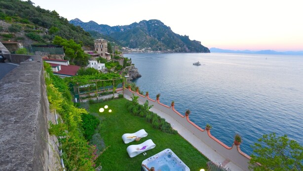 Area piscina con lettini e vasca idromassaggio, affacciata su un lago con montagne sullo sfondo.