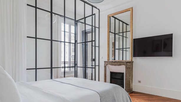 A bedroom with a bed, a large mirror, a fireplace, and a TV mounted on the wall.