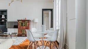 A dining area with a round table and white chairs, a wooden cabinet with decorative elements, and a television on a shelf.