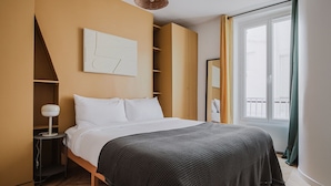 A modern bedroom with a bed, a bedside table, a lamp, a wall-mounted shelf, and a window with curtains.