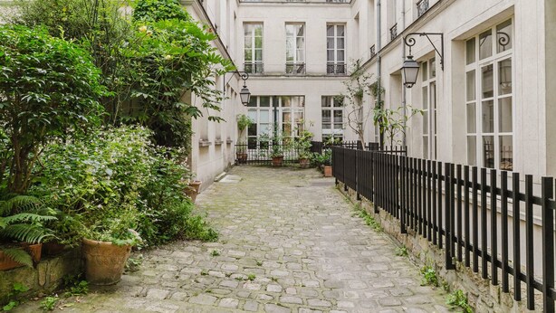 A narrow cobblestone courtyard with a black metal fence, surrounded by lush greenery and potted plants. - Blueground | Le Marais-Republique, nr restaurants (Paris)