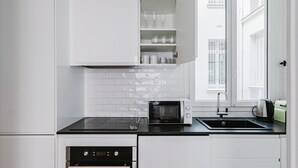 A modern kitchen with white cabinetry, a black countertop, a built-in microwave, a stainless steel sink, and a window with blinds. - Blueground | Porte Dauphine, elevator & washer (Paris)