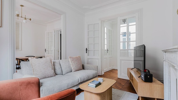 A modern living room with a grey sofa, a red armchair, a wooden coffee table, and a TV on a wooden stand. - Blueground | Porte Dauphine, elevator & washer (Paris)