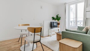 A modern, minimalist living room with a round dining table, two chairs, a green sofa, a TV on a white stand, and a window with a view.
