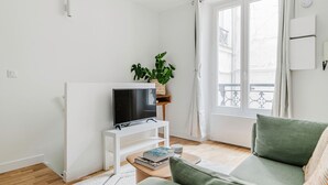 A living room with a flat-screen TV on a white stand, a green sofa, a small coffee table with books, a potted plant, and a window with a view of a balcony.
