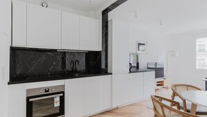 Et moderne køkken med hvide skabe, en backsplash i sort marmor og en indbygget ovn. Der er et spiseområde med et rundt bord og stole.