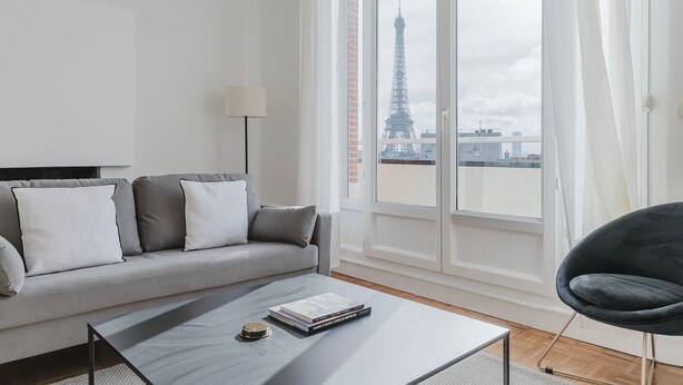 Un moderno salón con un sofá gris, una silla negra y una mesa de centro. Un ventanal ofrece vistas a la Torre Eiffel.