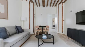 Ein modernes Wohnzimmer mit grauem Sofa, einem schwarzen Couchtisch und einem Esstisch mit Stühlen. Ein Fernseher ist an der Wand montiert und es gibt eine Holzbalkendecke.