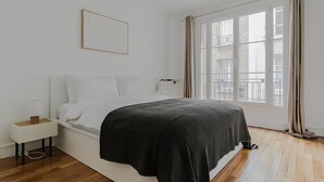 A bedroom with a bed, a nightstand, a radiator, and a window with curtains. - Blueground | Batignolles 2bd apartment (Paris)