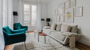 A modern living room with a teal armchair, a beige sofa, and a white rug. There are framed artworks on the wall and a floor lamp. - Blueground | Batignolles 2bd apartment (Paris)