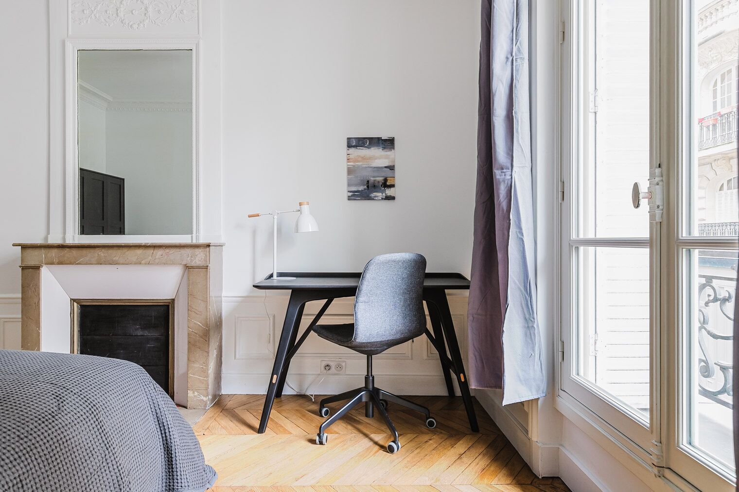 Een slaapkamer met een bed, een bureau met stoel, een spiegel, een open haard en een raam met gordijnen.
