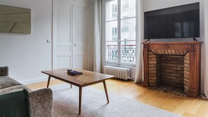 A living room with a fireplace, a television mounted above it, a window with a view of buildings, and a door. - Blueground | Notre-Dame-de-Lorette 1bd apartment (Paris)