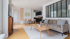 A modern living room with a sofa, coffee table, and dining area. There is a kitchen in the background with a large window. - Blueground | Notre-Dame-de-Lorette 1bd apartment (Paris)