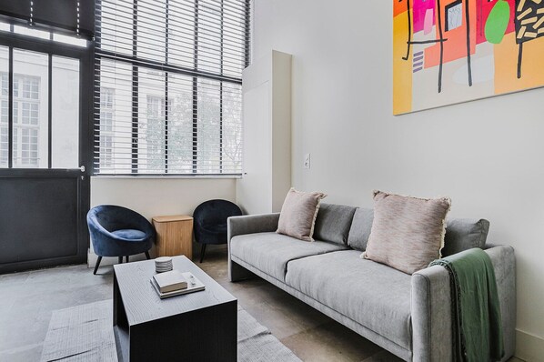 Un salon moderne avec un canapé gris, deux fauteuils, une table basse et une grande fenêtre équipée de stores.