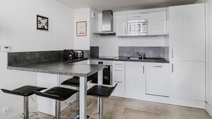 Una cocina moderna con armarios blancos, encimera de acero inoxidable y microondas integrado. Hay dos taburetes de bar y una cafetera.