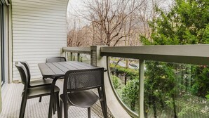 Un balcon avec une table et des chaises, une vue sur des arbres et un étang, et une maison aux murs recouverts de bardage blanc.