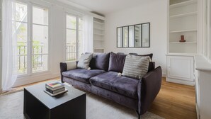 A modern living room with a dark purple sofa, a coffee table with books, and a large window with curtains. - Blueground | Le Marais, elevator & w/d, nr Seine (Paris)