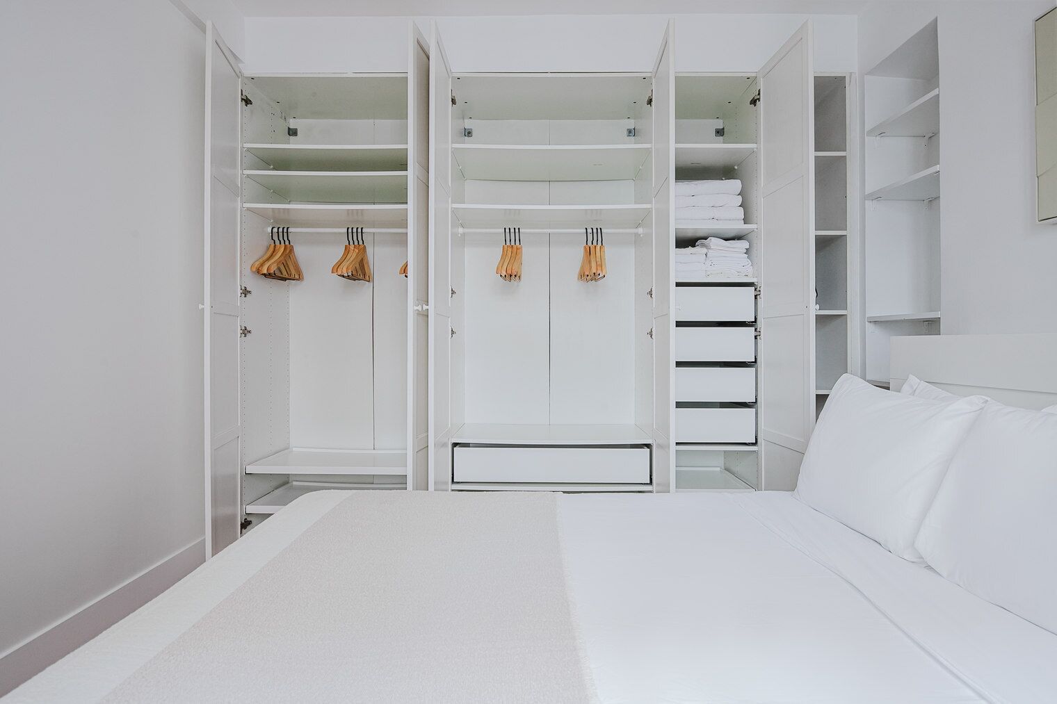 A bedroom with a white bed, a large wardrobe with multiple shelves, and a bedside table.