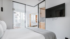 Habitación de hotel moderna con una cama grande, televisor montado en la pared y puerta corrediza de vidrio que da a un balcón con zona de comedor.
