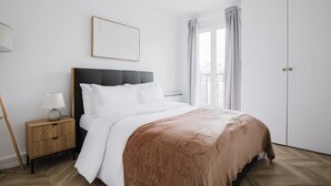 Un dormitorio con cama, mesita de noche, lámpara, ventana con cortinas y puerta.