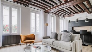 A modern living room with a sofa, a glass coffee table, and a chair. There are large windows with curtains, a radiator, and a kitchen area with a countertop and cabinets. - Blueground | Odéon, washer, nr Luxembourg Gdns (Paris)