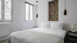 A modern bedroom with a large bed, two bedside tables, a wall-mounted artwork, and a window with a view of buildings.