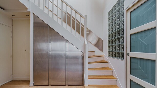 A staircase with wooden steps and white railings leading to a room with a glass door and a metal closet. - Blueground | Le Marais, elevator & w/d, nr Vosges (Paris)