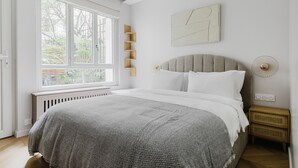 A bedroom with a large window, a bed with a grey and white patterned bedspread, a grey upholstered headboard, and a wall-mounted light fixture. - Blueground | Auteuil, washer, nr Monceau Park (Paris)