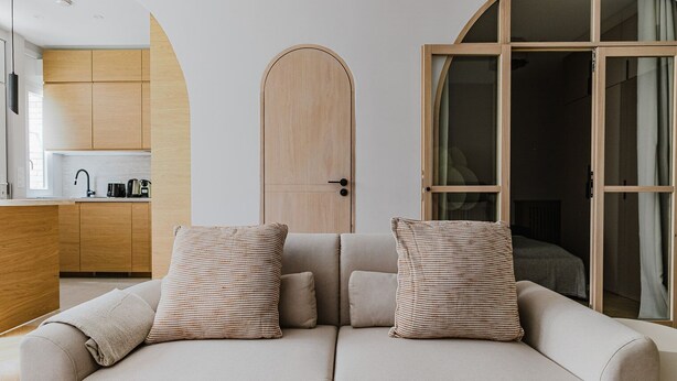 A modern living room with a sofa, two patterned pillows, a wooden door, and a view into a bedroom. - Blueground | Auteuil, washer, nr Monceau Park (Paris)