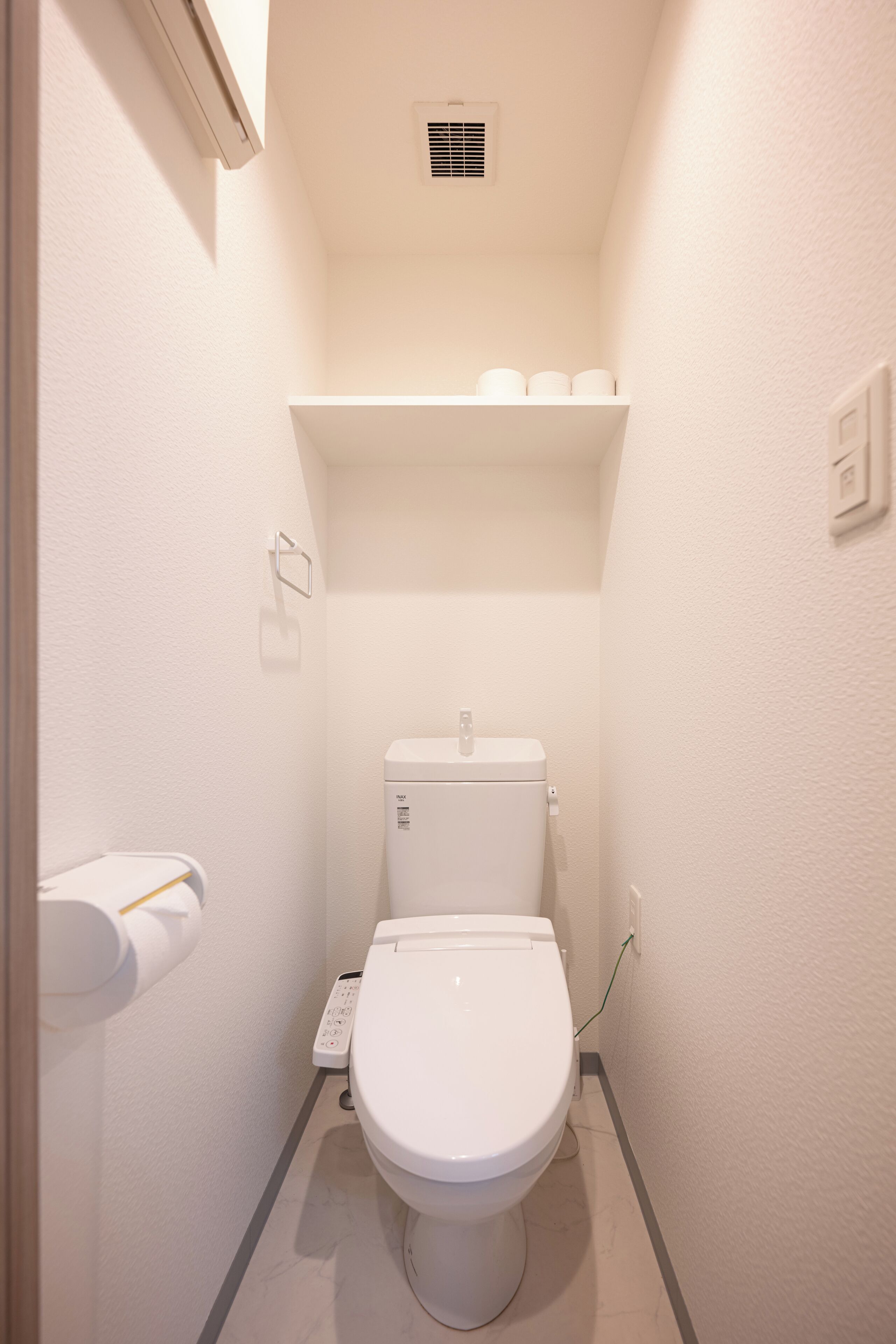 A compact, modern bathroom with a white toilet, a shelf above the toilet, and a wall-mounted vent.