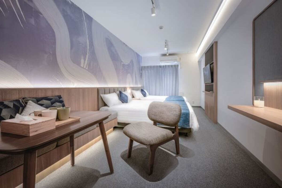 A modern hotel room with a bed, a desk with a chair, a large abstract artwork on the wall, and a television on the right.