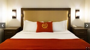 Una camera d'albergo con un letto grande, una coperta rossa e un cuscino con un logo. Sono presenti due comodini, con lampade e un telefono su uno di essi.
