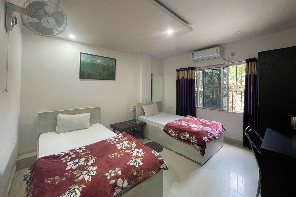 A hotel room with two beds, a desk, a chair, a fan, a window with curtains, and an air conditioning unit.