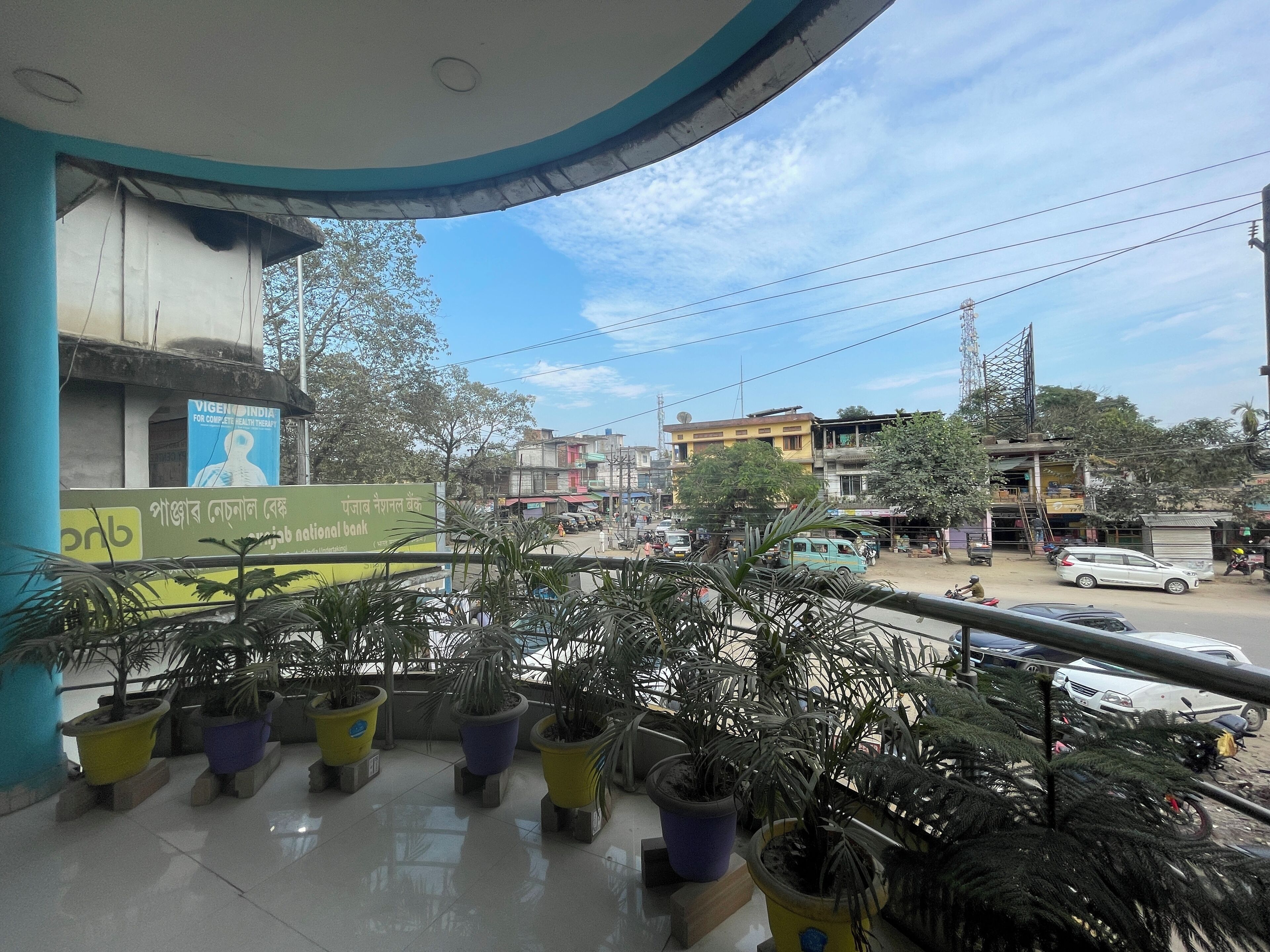 Un balcón con plantas en macetas, un letrero de banco y vista a una calle concurrida con vehículos y edificios.