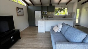 Una cocina moderna con microondas integrado, refrigerador y pileta. Hay un sofá gris con un cojín decorativo y un televisor de pantalla plana sobre un pedestal negro. La habitación tiene vigas de madera en el techo y ventanas amplias.