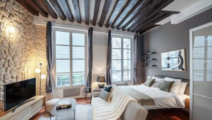 A modern bedroom with a stone wall, a large bed, a TV, and a cozy seating area. - Blueground | Le Marais, near Seine River (Paris)