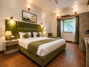 A hotel room with a large bed, two bedside tables, a nightstand, a green upholstered headboard, a painting on the wall, a window with curtains, and a marble accent wall. - Staybook The Rudveda Resort (Ramnagar)